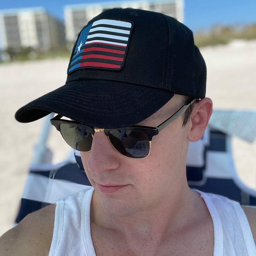 Lifestyle image of a man wearing the hat and sunglasses sitting at the beach.